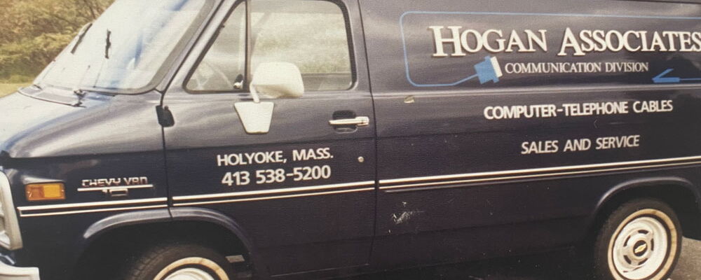 A vintage blue Chevrolet van with "Hogan Associates Communication Division" branding on the side, parked outdoors. The van features the company details, including "Holyoke, Mass." and a phone number, alongside text advertising "Computer-Telephone Cables,