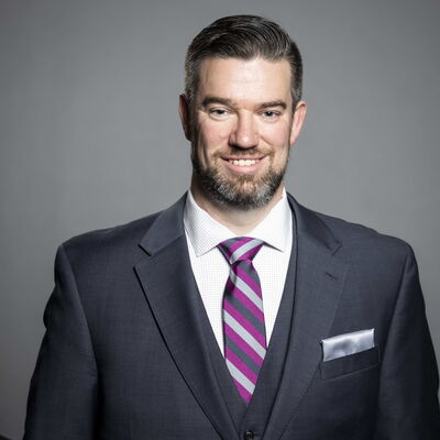 Man in a tailored suit with striped tie and pocket square, smiling confidently.
