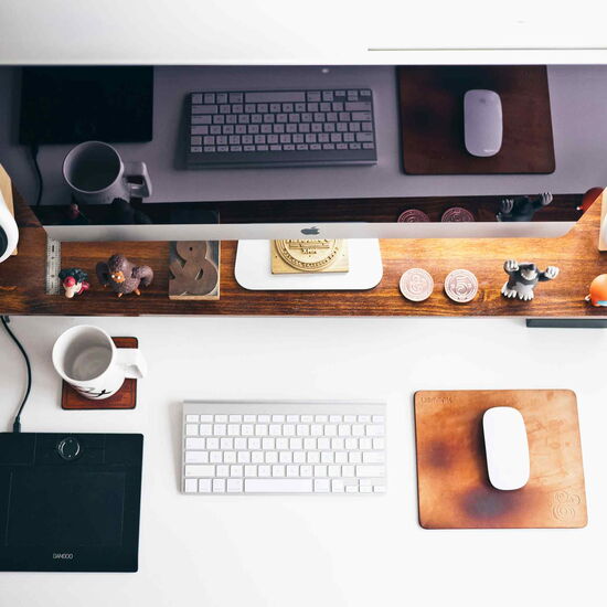 A well-organized desk setup featuring an Apple iMac monitor, a wooden shelf with small figurines and coins, two white speakers, and a keyboard and mouse on the desk. Additional items include a drawing tablet, a coffee mug, and a leather mouse pad, creatin