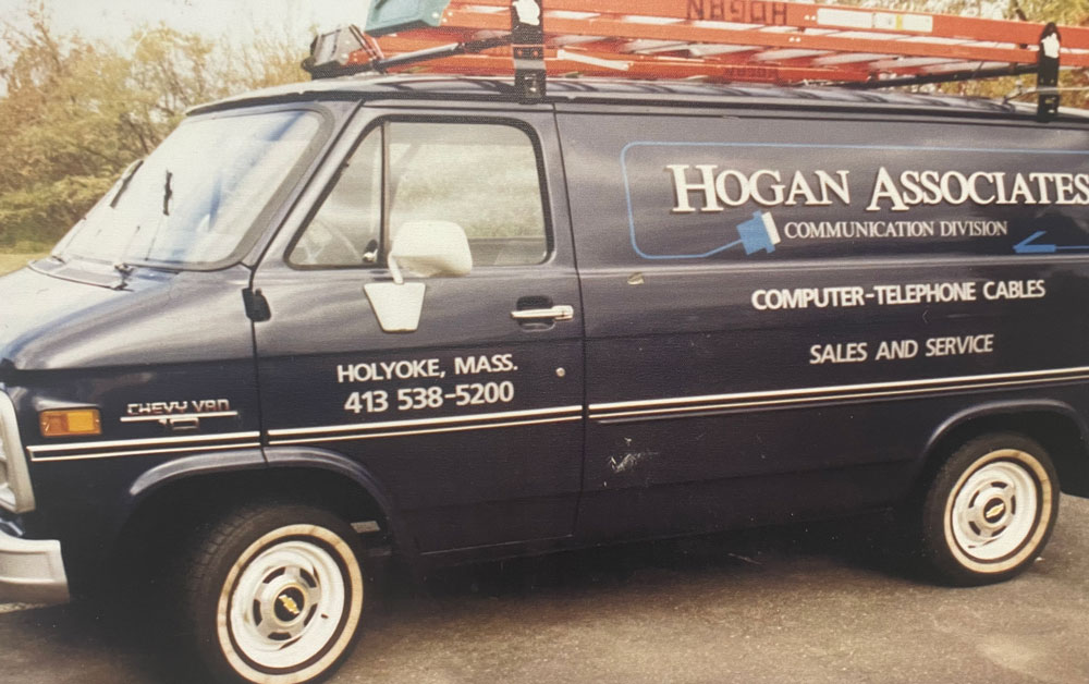 A vintage blue Chevrolet van with "Hogan Associates Communication Division" branding on the side, parked outdoors. The van features the company details, including "Holyoke, Mass." and a phone number, alongside text advertising "Computer-Telephone Cables,