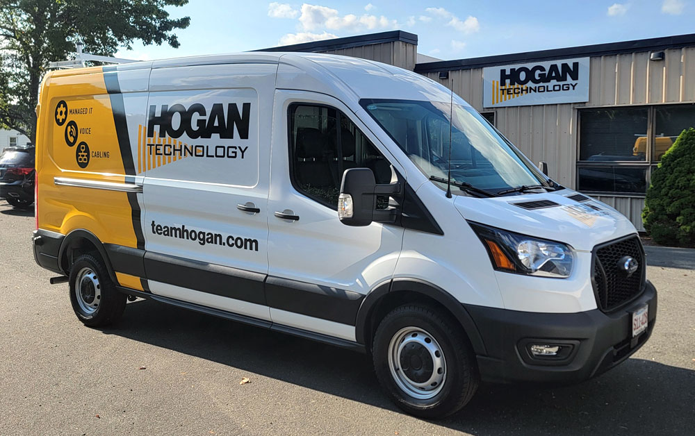 A white and yellow Ford van branded with "Hogan Technology" and "teamhogan.com," featuring icons for Managed IT, Voice, and Cabling services. The van is parked in front of a Hogan Technology building, which has the company logo displayed prominently on th