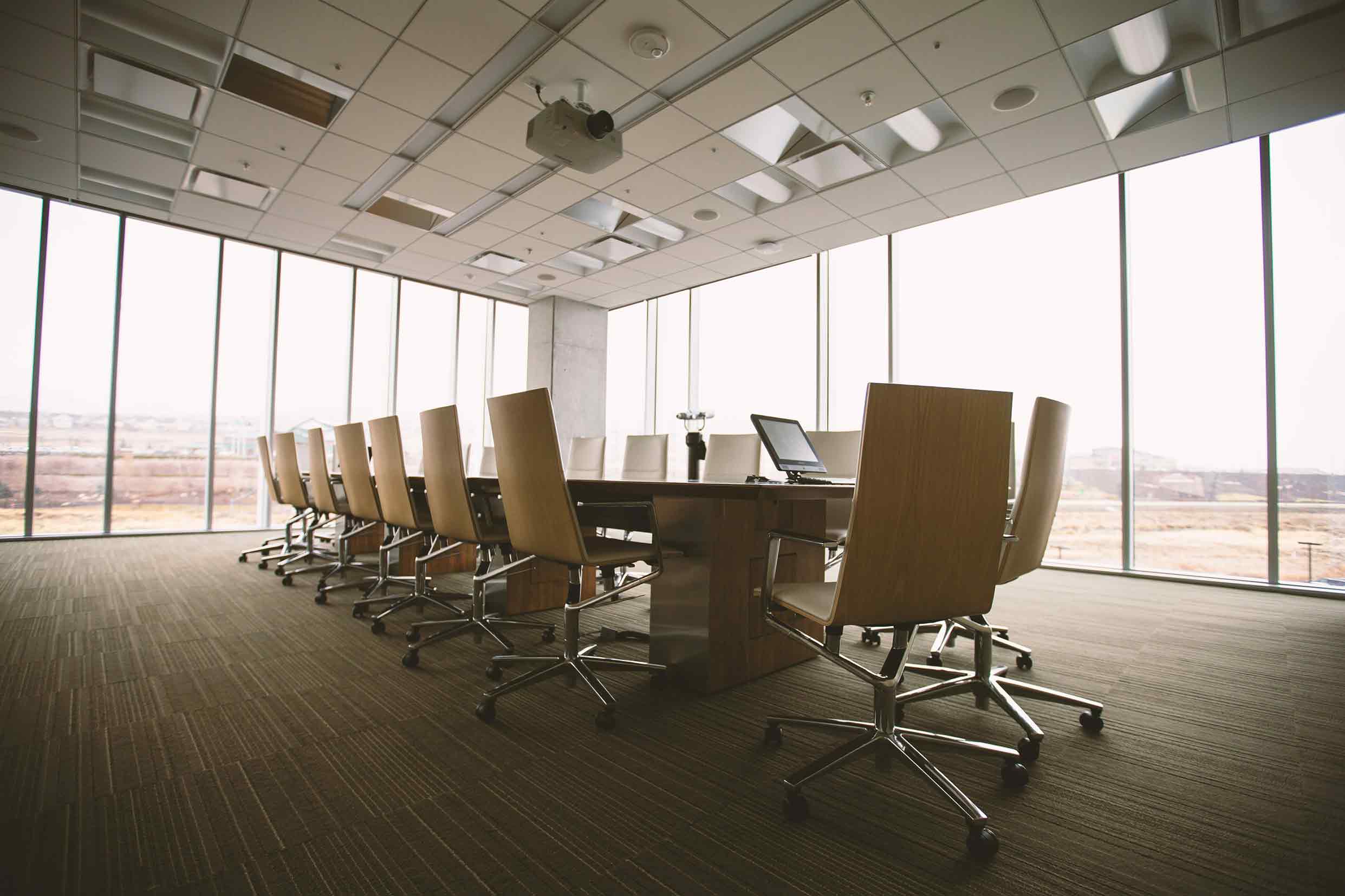 A modern conference room with a long wooden table surrounded by rolling office chairs. The room features floor-to-ceiling windows offering a view of an outdoor landscape, and a ceiling-mounted projector. The space is bright and professional, ideal for mee