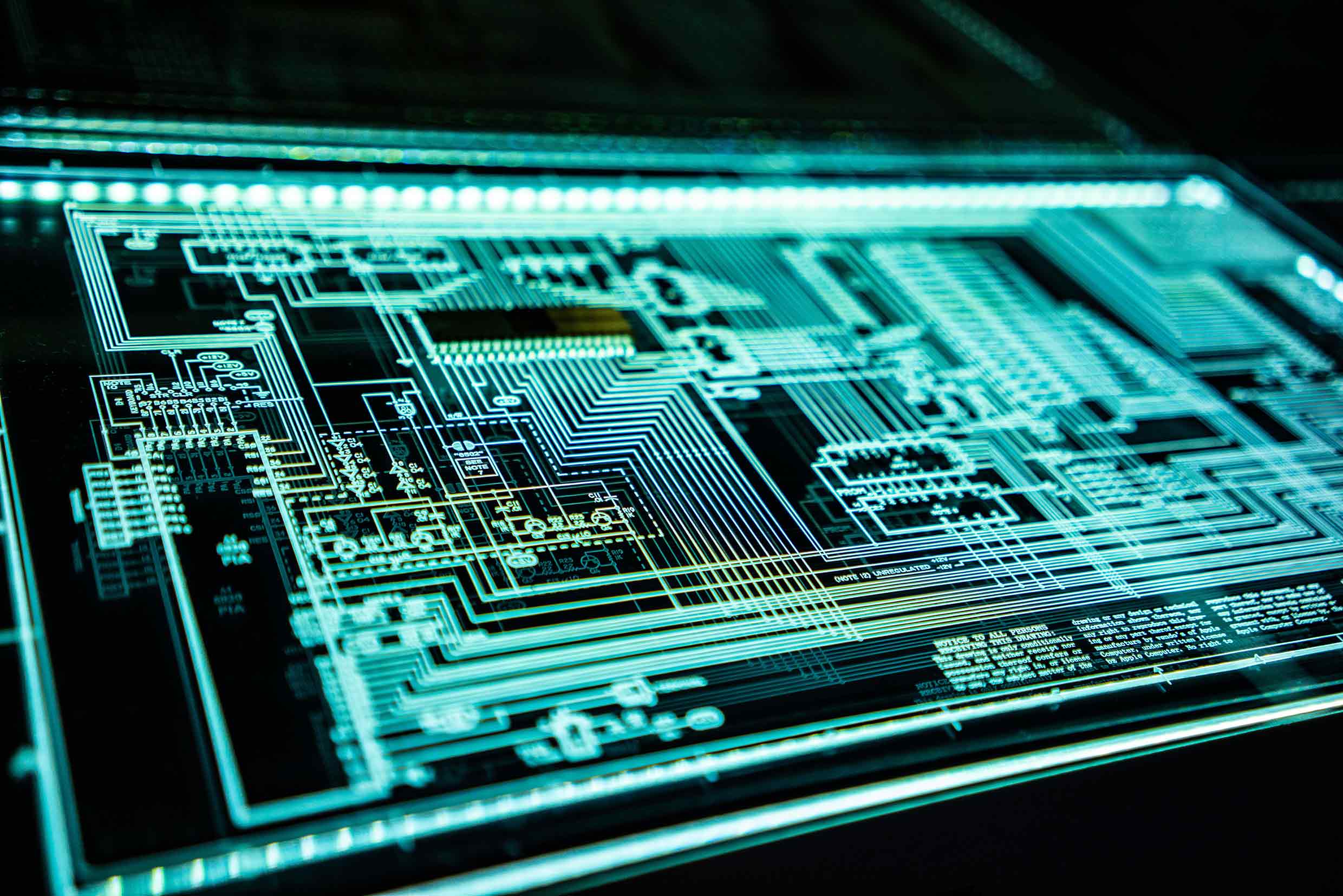 A glowing digital circuit board design displayed on a high-tech screen, featuring intricate pathways, nodes, and components. The image highlights the complexity of electronic systems and technology, with a futuristic and sleek appearance.