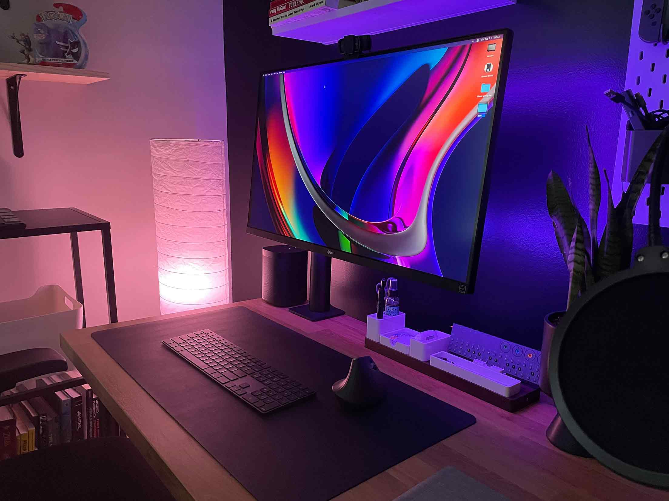 A modern and stylish desk setup featuring a widescreen monitor displaying a vibrant abstract design, illuminated by purple ambient lighting. The desk includes a keyboard, a vertical ergonomic mouse, and organized accessories. A soft-glow floor lamp and a