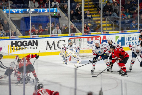 A live hockey game in action with players from opposing teams battling near the goal. The goalie, dressed in colorful pads, is positioned to defend. The arena features advertising boards with logos, including "Hogan Technology." Spectators fill the stands